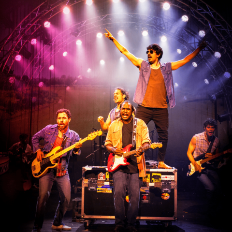 Five musicians/actors on-stage, two with electric guitars, one on drums and one with arms in air, under pink and purple spotlights