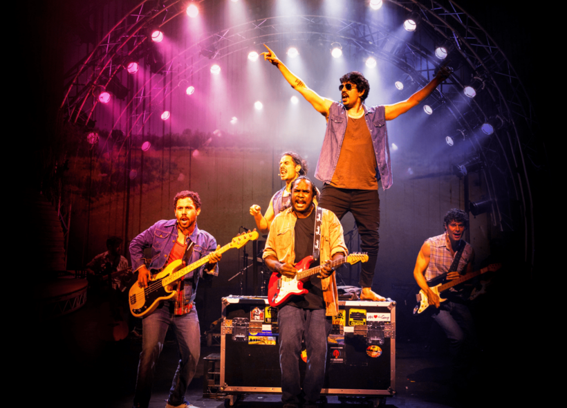 Five musicians/actors on-stage, two with electric guitars, one on drums and one with arms in air, under pink and purple spotlights