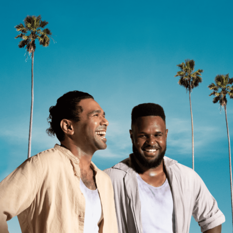 Two Torres Strait Islander men in neutral linen shirts stand smiling; one looking to the right, one straight at the camera, blue sky backdrop with 3 tall and skinny palm trees