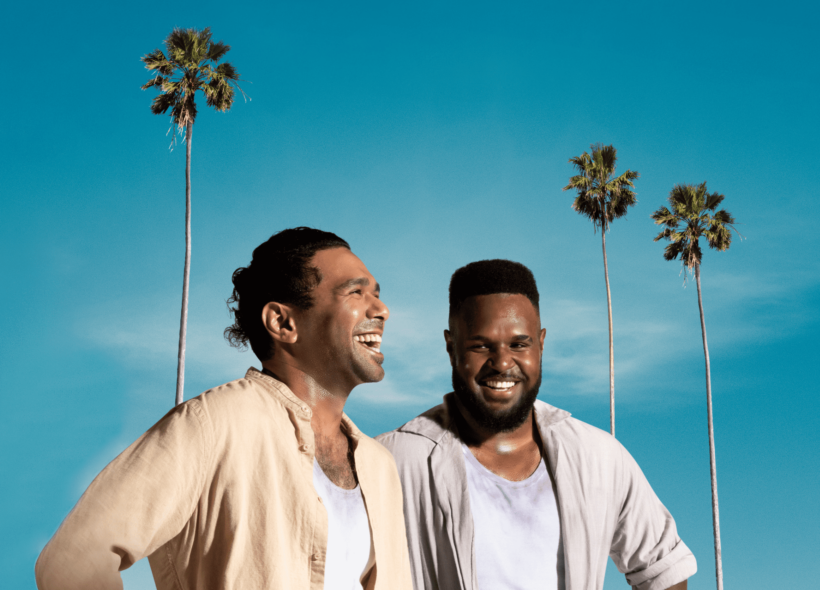 Two Torres Strait Islander men in neutral linen shirts stand smiling; one looking to the right, one straight at the camera, blue sky backdrop with 3 tall and skinny palm trees