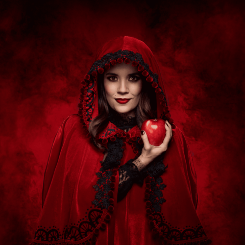 A woman dressed as Red Riding Hood in red cape, wearing red lipstick, holding a red apple with a sinister red background