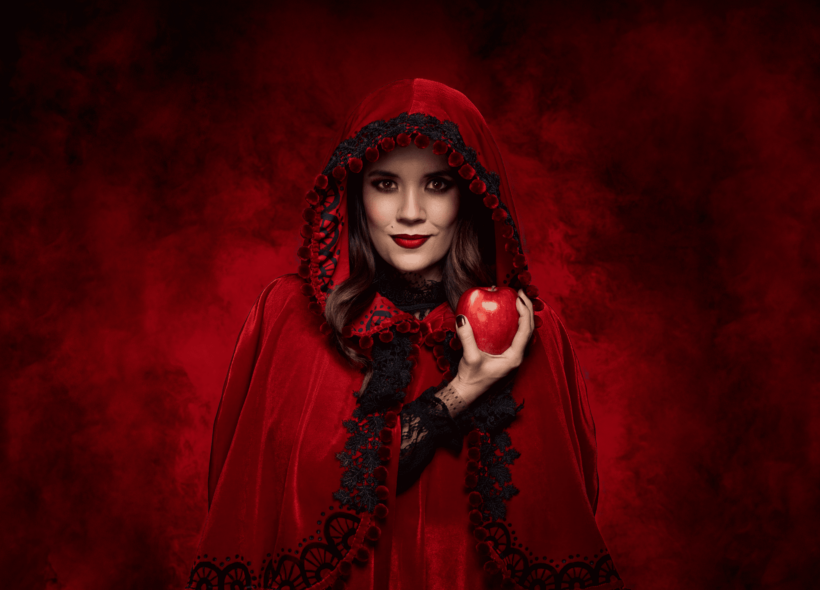 A woman dressed as Red Riding Hood in red cape, wearing red lipstick, holding a red apple with a sinister red background