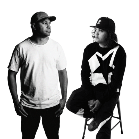 A black and white still photo of Two First Nations men, the lead artists, one standing with hands in pockets and one sitting on a stool