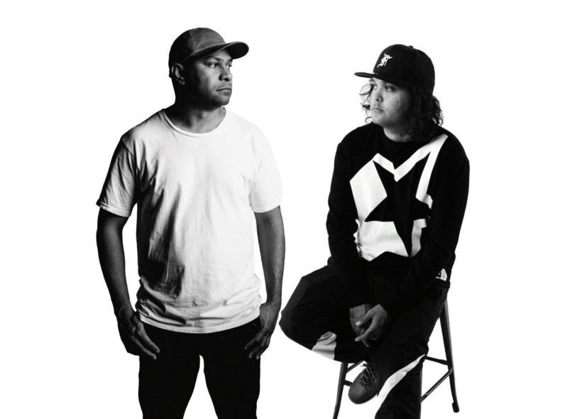 A black and white still photo of Two First Nations men, the lead artists, one standing with hands in pockets and one sitting on a stool