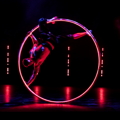 Shirtless male acrobat/dancer suspended in huge metal ring on a darkened stage highlighted with small red lights