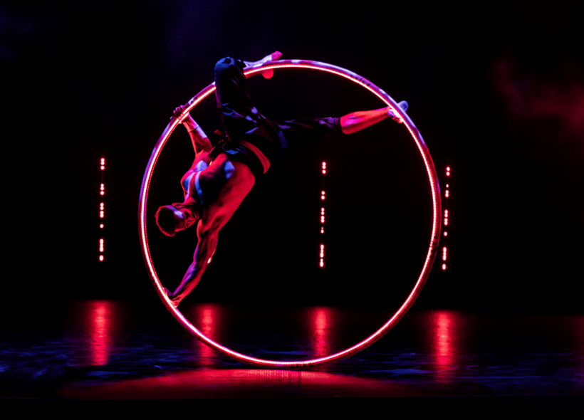 Shirtless male acrobat/dancer suspended in huge metal ring on a darkened stage highlighted with small red lights