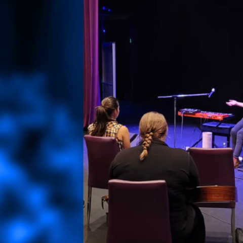 A lady sitting on a chair on a stage, facing the audience. There is 2 microphones and a keyboard on the stage next to here. A blue overlay is placed on top of the photo.