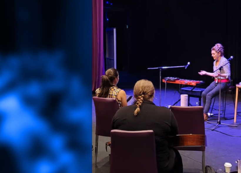 A lady sitting on a chair on a stage, facing the audience. There is 2 microphones and a keyboard on the stage next to here. A blue overlay is placed on top of the photo.