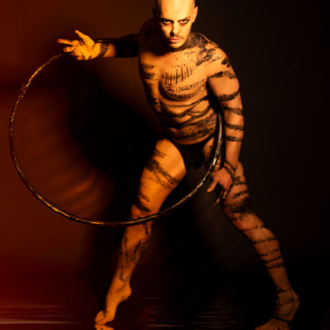 A man wearing only black underwear and who's body is covered in black paint streaks appears to be standing on water. The man looks just below the camera lense and is holding a black hoola-hoop over his waist and shoulders. His body leans into the camera and his facial expression holds a serious, dark and contemplative expression.