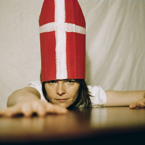 Rose Maher in a white t-shirt and cardinal's mitre reaches across a table