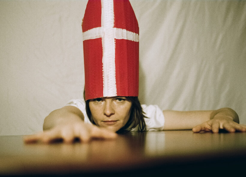 Rose Maher in a white t-shirt and cardinal's mitre reaches across a table