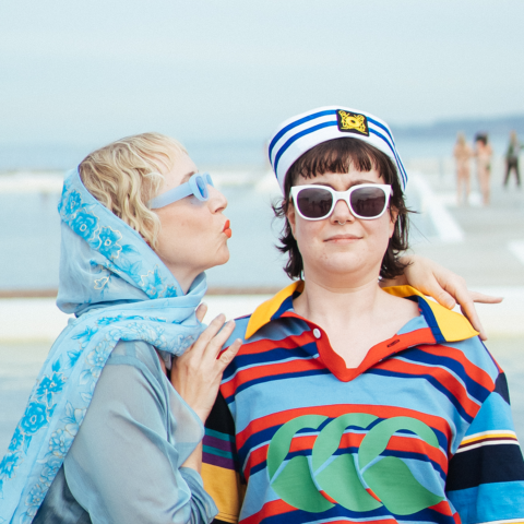 Two actors stand in front of the Newcastle Ocean Baths. One is about to kiss the other on the cheek.