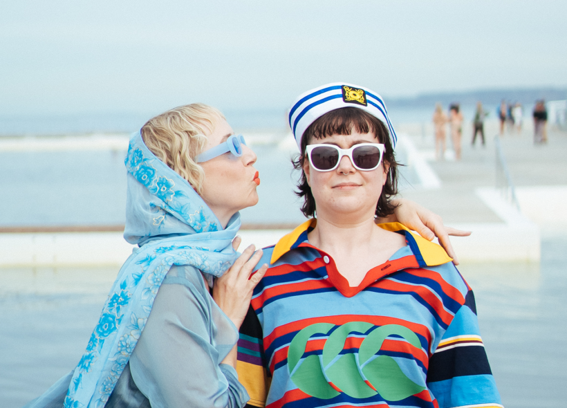 Two actors stand in front of the Newcastle Ocean Baths. One is about to kiss the other on the cheek.