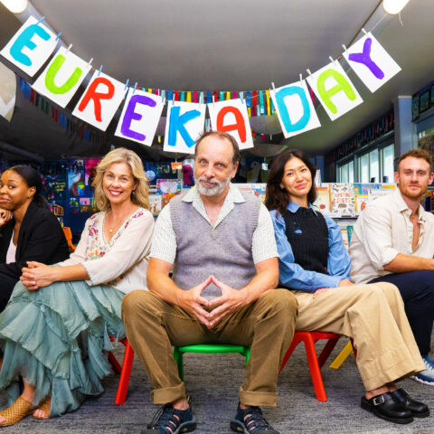 The cast of Eureka Day sit on children stairs in a classroom. 