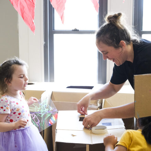 Cubbyhood production photo: Suzanne Phoenix. A Polyglot artist and two children create and play amongst cardboard box cubbies with masking tape, acetate and pastels. Red paper bunting hangs above them.