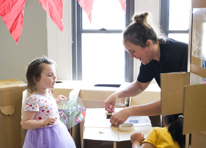 Cubbyhood production photo: Suzanne Phoenix. A Polyglot artist and two children create and play amongst cardboard box cubbies with masking tape, acetate and pastels. Red paper bunting hangs above them.