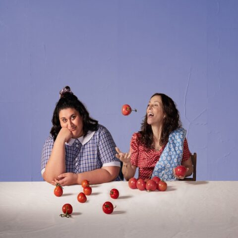2 women in front of purple background throwing tomatoes in air