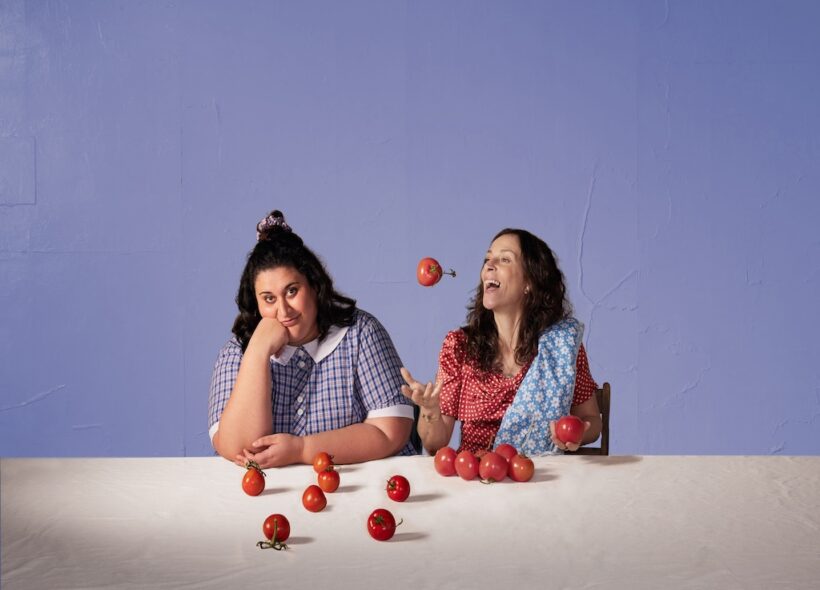 2 women in front of purple background throwing tomatoes in air