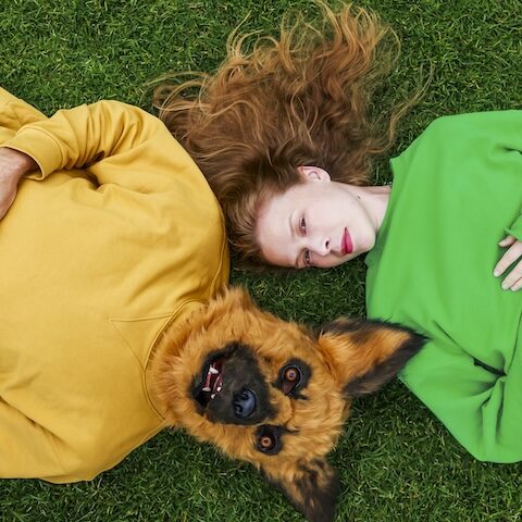 Lucy Heffernan lays in a green jumpy on grass, with a man in a yellow jumper with a dog head lays to her left