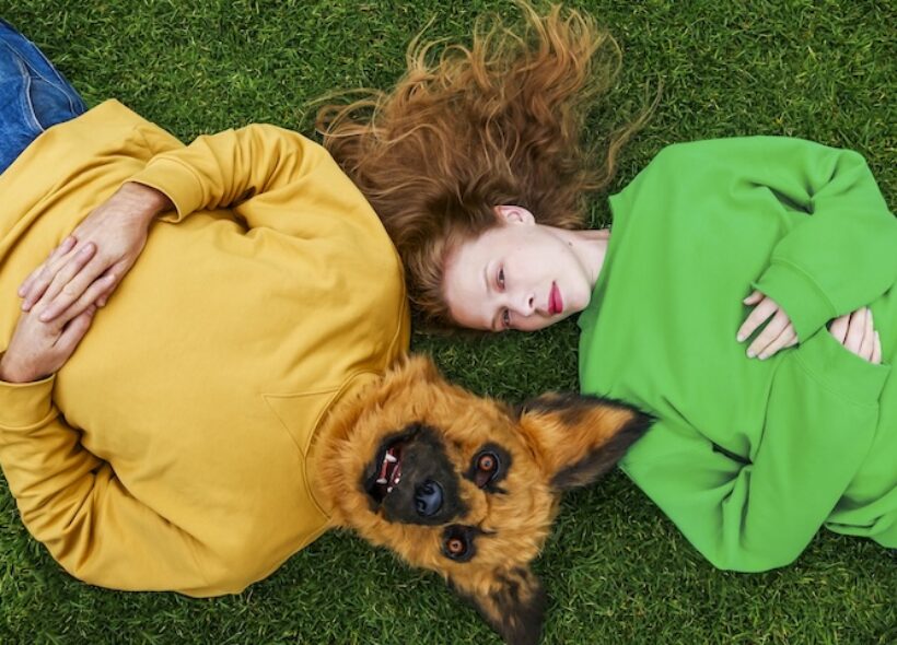 Lucy Heffernan lays in a green jumpy on grass, with a man in a yellow jumper with a dog head lays to her left