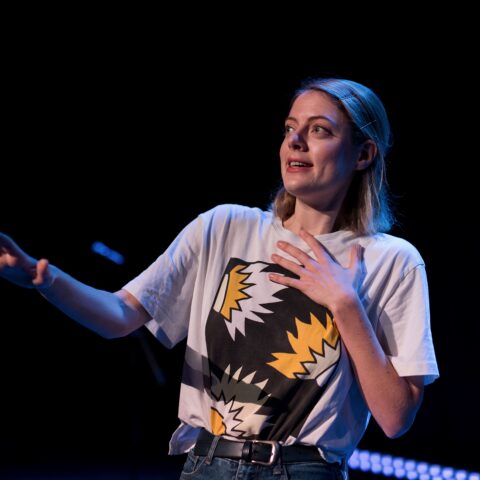woman with blonde hair wearing a white tshirt with a colourful print on the front in front of a black background