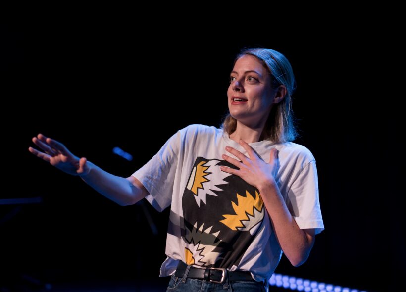 woman with blonde hair wearing a white tshirt with a colourful print on the front in front of a black background