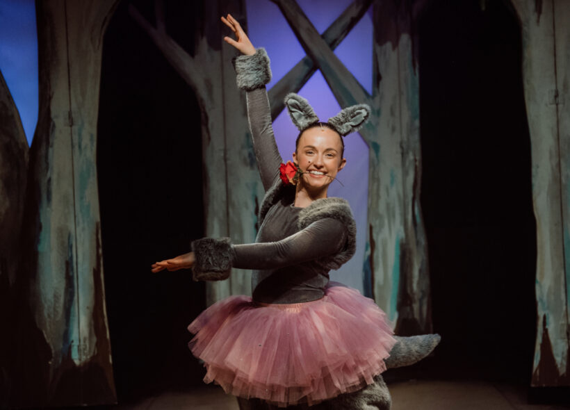 Young female actress in a pink tutu and kangaroo ears posing with a red rose in her teeth in a ballet pose. 