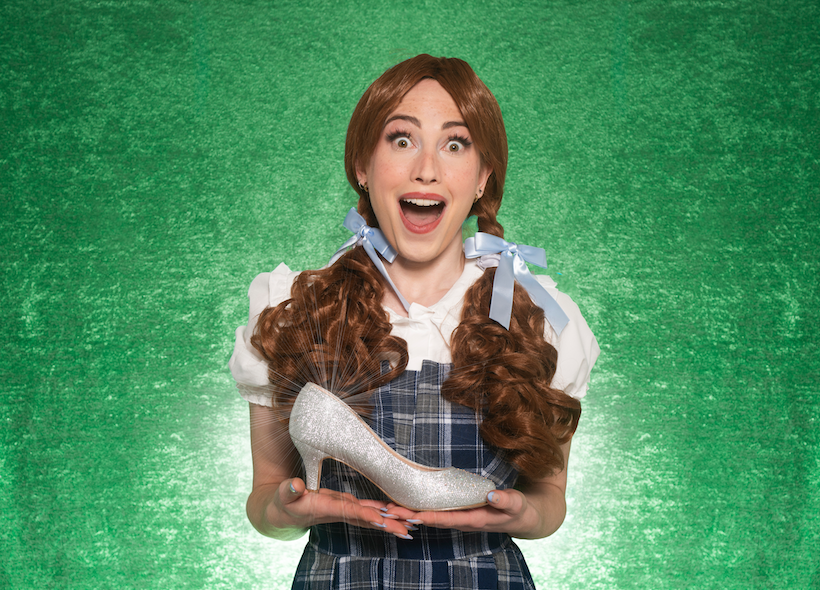 Joyfully smiling woman in low pigtails and checkered dress holding a silver slipper agains an emerald background