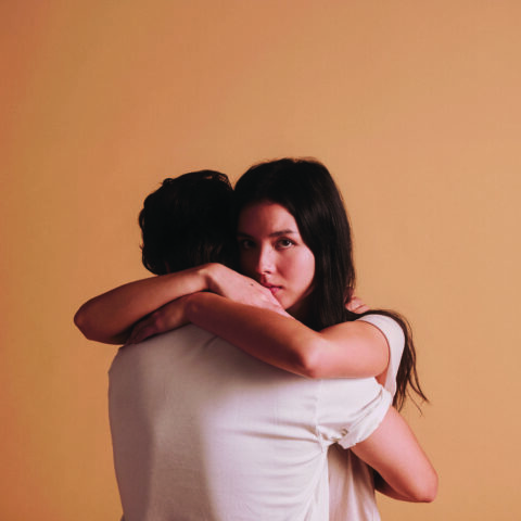 Two teenagers embracing, Juliet looks intensely over Romeo's shoulder. Plain apricot background. 