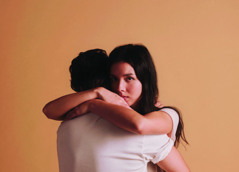 Two teenagers embracing, Juliet looks intensely over Romeo's shoulder. Plain apricot background. 