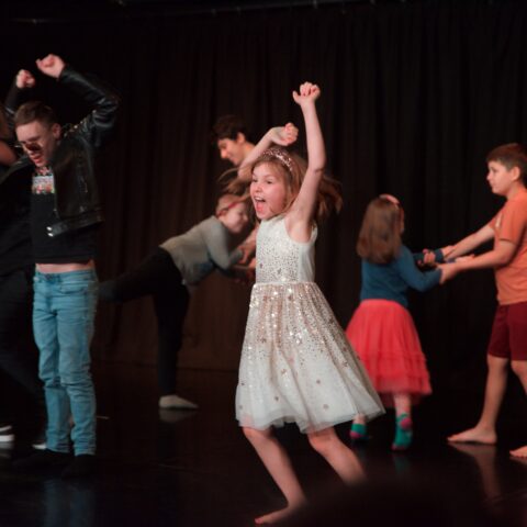 Children perform during a Restless Dance Theatre end-of-year showing. A child in a sparkly white dress dances energetically in the foreground while other children move and play together on stage.