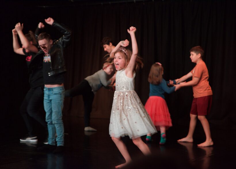Children perform during a Restless Dance Theatre end-of-year showing. A child in a sparkly white dress dances energetically in the foreground while other children move and play together on stage.