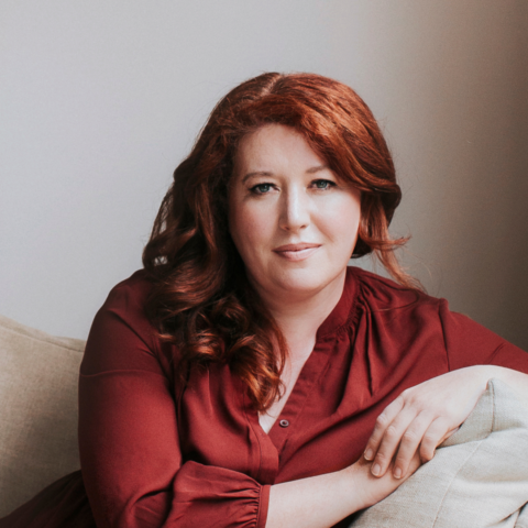 Author Jane Harper sits on a beige sofa, leaning over the arm, looking directly at the camera. 
