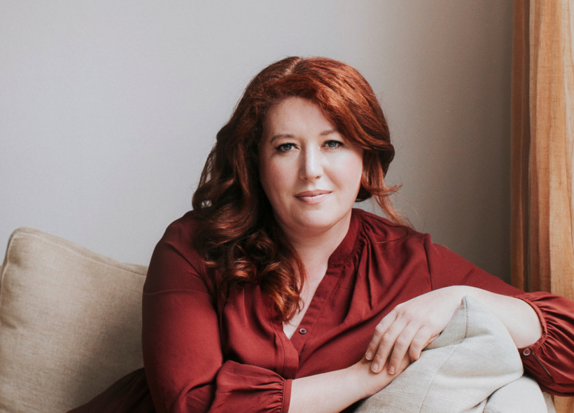 Author Jane Harper sits on a beige sofa, leaning over the arm, looking directly at the camera. 