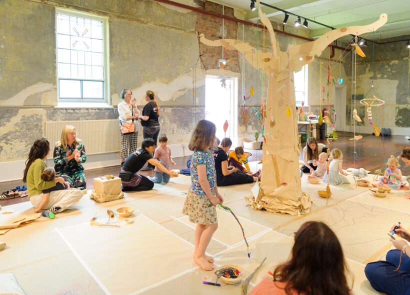 Art Tree production photo: Sarah Walker. A child in blue stands in the middle of the room holding a stick. The cardboard art tree is centre frame. Families and Polyglot artists are scattered around the room in the background.