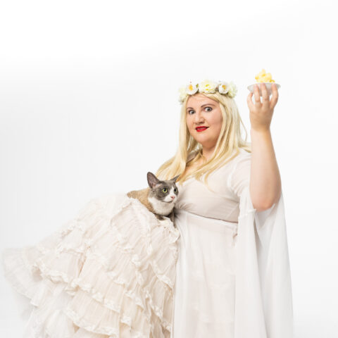 Cousin Tara is sitting, wearing a white ruffled gown and petticoat, a blonde wig and flower crown. She holds a small dish of butter in the air with her left hand, with a grey and brown cat on her lap looking at the butter. She is looking intensely at the camera in the centre of the frame.