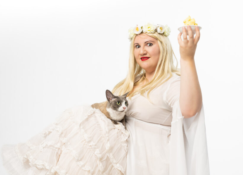 Cousin Tara is sitting, wearing a white ruffled gown and petticoat, a blonde wig and flower crown. She holds a small dish of butter in the air with her left hand, with a grey and brown cat on her lap looking at the butter. She is looking intensely at the camera in the centre of the frame.