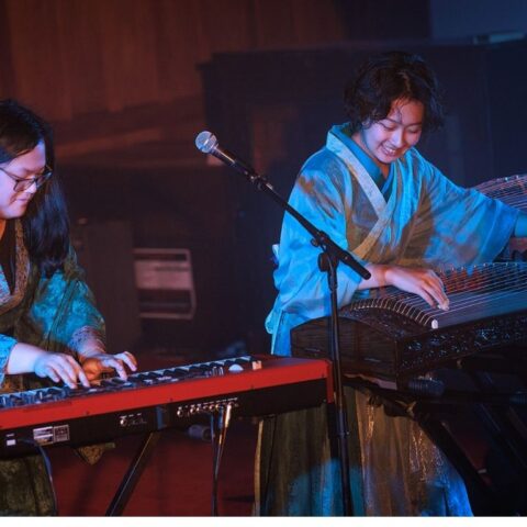 Two musicians perform on stage: one playing a red keyboard, the other playing the guzheng, both wearing traditional-style robes.