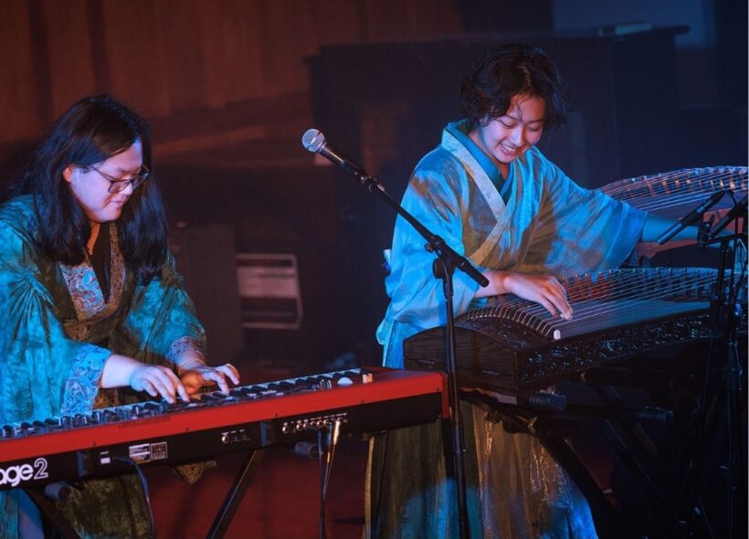 Two musicians perform on stage: one playing a red keyboard, the other playing the guzheng, both wearing traditional-style robes.