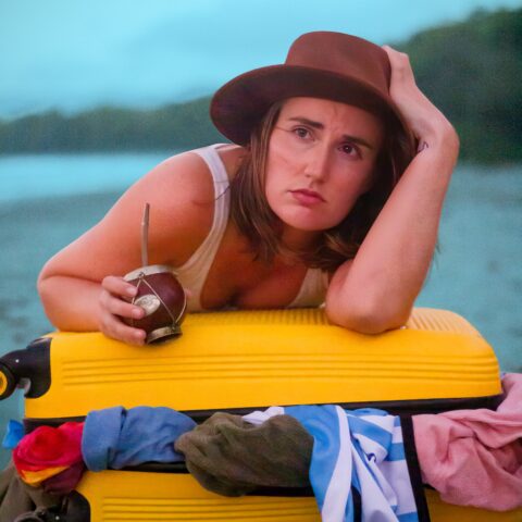 A woman leaning on a open suitcase, holding a mate and wearing a hat.