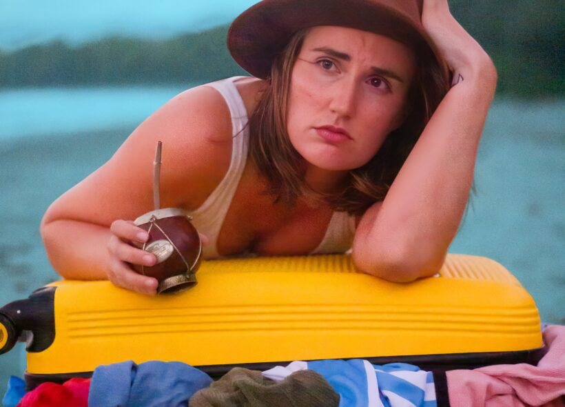 A woman leaning on a open suitcase, holding a mate and wearing a hat.