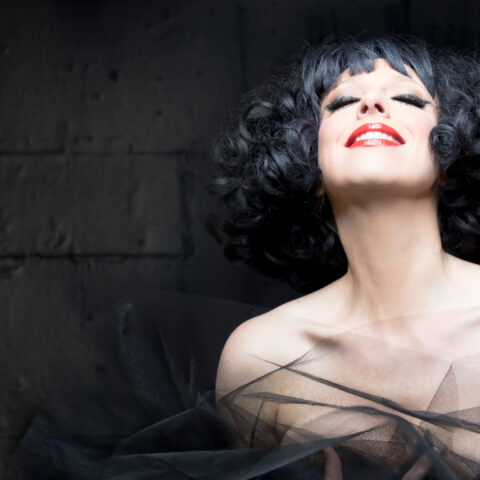 A woman with black curly hair and striking red lipstick, smiling softly against a grey background