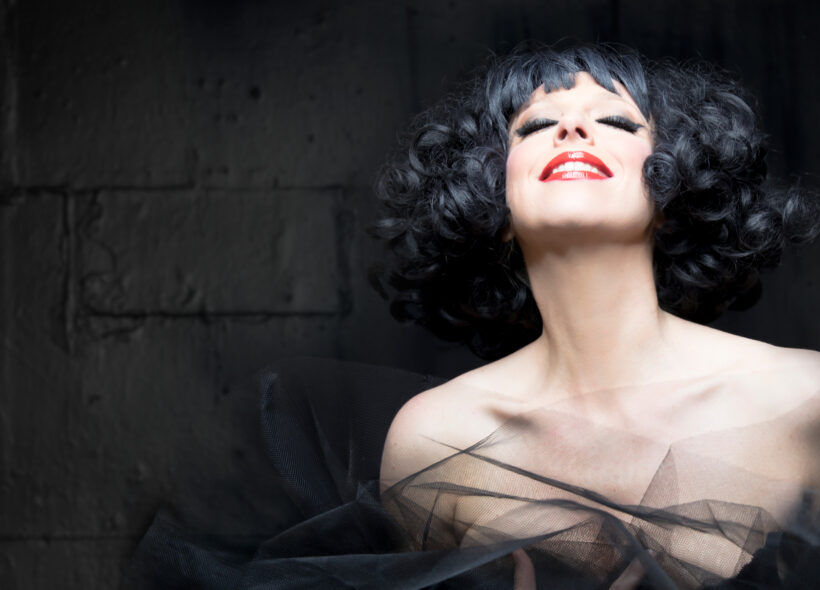 A woman with black curly hair and striking red lipstick, smiling softly against a grey background