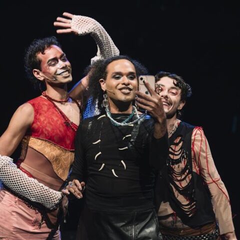Photograph of the 'Motherless Collective' First Nations Drag artists performing their theatre show '3 Blak Ravers' the background is plain black, Stone Motherless cold is standing on the left, with their arm up, Cerulean is in centre, holding a phone up. On the right is Mora Money. They are all smiling. 