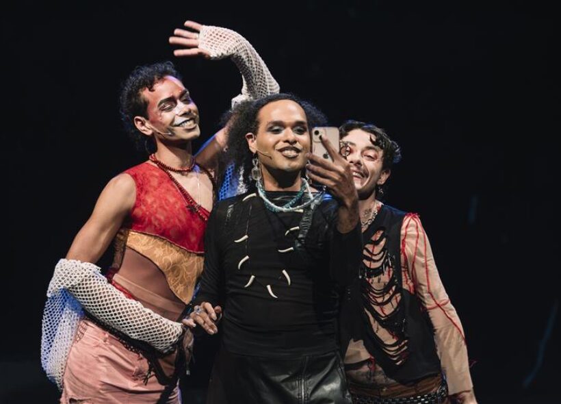 Photograph of the 'Motherless Collective' First Nations Drag artists performing their theatre show '3 Blak Ravers' the background is plain black, Stone Motherless cold is standing on the left, with their arm up, Cerulean is in centre, holding a phone up. On the right is Mora Money. They are all smiling. 