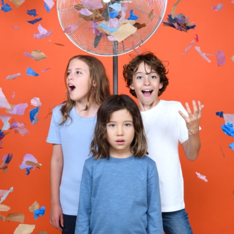 Three children stand against an orange wall, in-front of a fan. There is confetti in the air. Photographer: Sarah Walker.