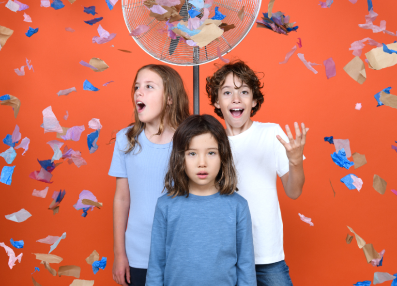 Three children stand against an orange wall, in-front of a fan. There is confetti in the air. Photographer: Sarah Walker.