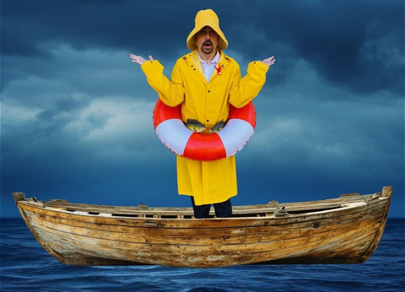 Jeremy Moses stands with arms up in a confused pose, in an old wooden boat in the ocean. He wears a yellow long raincoat with black suit pants, with a red and white lifesaving buoy around his waist. He is modestly covered in small crabs and fish.
