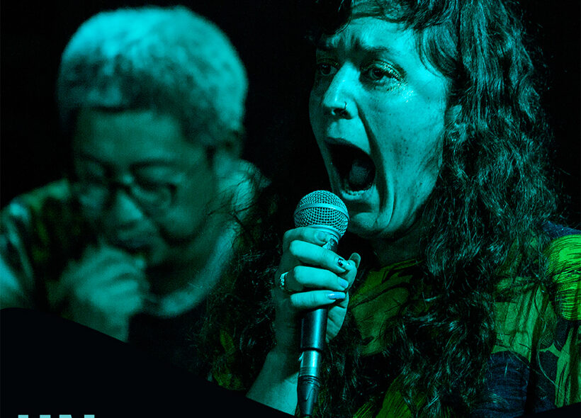 In the foreground, a white woman holds a microphone with her mouth gaping wide open. She has long wavy brown hair. A loose dress that has shades of navy with large yellow flowers. Nail polish and glitter around her eyes. In the background to the left is a Korean man with glasses, greyish short hair blowing into a small nondescript instrument.