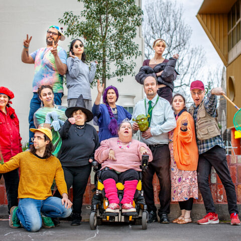 12 colourfully dressed people stand, sit and kneel around a low brick wall. Each have larger than life expressions on their faces. In the background are trees and buildings